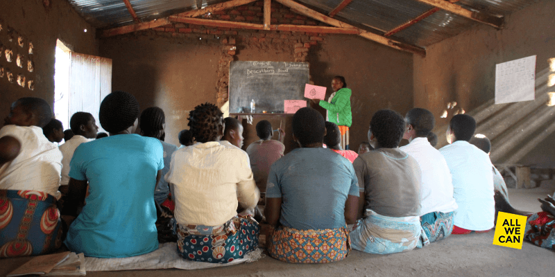 A teacher wearing a green jacket points to a blackboard in a classroom in Malawi. A group of girls watch attentively, their backs to the camera.